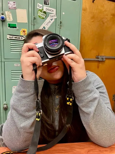 A student raising a 35mm film camera to her eye to take a photograph, sitting in front of a bank of green lockers covered in stickers