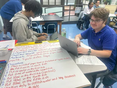 Two students working at laptops behind a large handwritten lab plan setting out the question, variables, materials and procedure for an experiment on how water affects cucumber seed germination