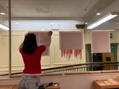 A student on a ladder hanging a large pale pink paper work on a wall for an exhibition, with two more already installed alongside