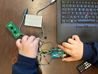 A student's hands wiring an Arduino board and breadboard next to a laptop, with an LCD module and jumper wires spread across the desk
