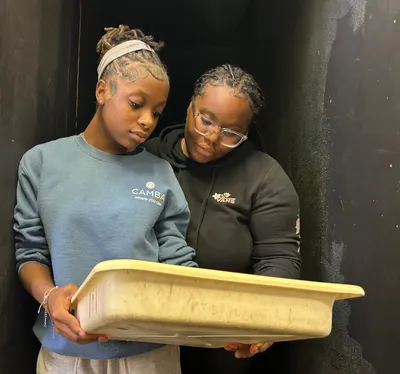 Two students standing in a darkroom, looking down at a developing tray they are holding between them