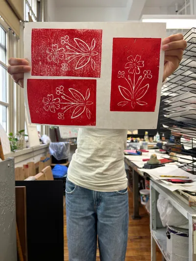 A student holding up a finished sheet of red block prints of flowers in the printmaking studio, drying racks behind them