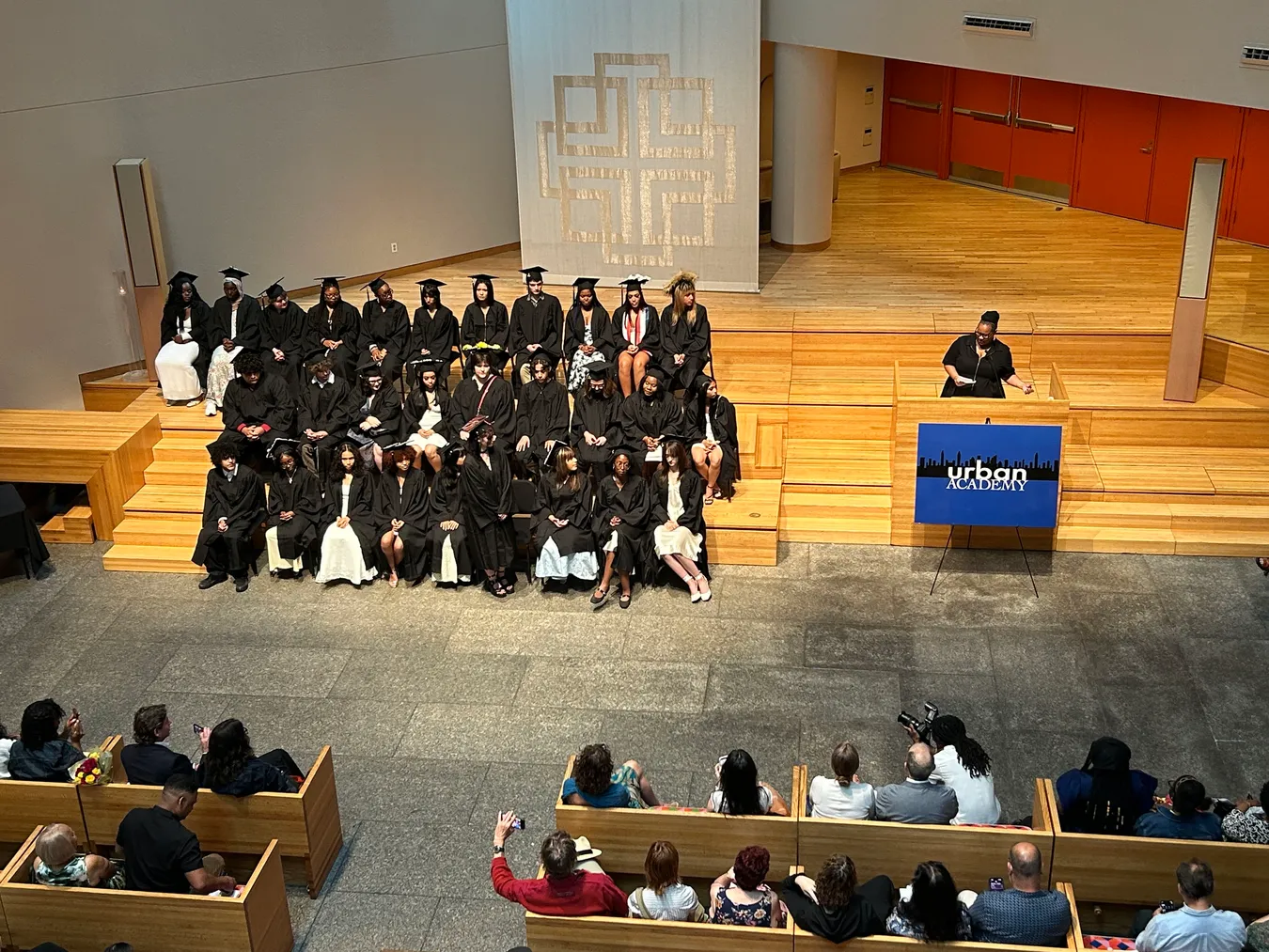 Graduating students in caps and gowns seated together on tiered wooden steps at a graduation ceremony, viewed from a balcony, with a speaker at a podium beside an Urban Academy sign and families watching from the seats below