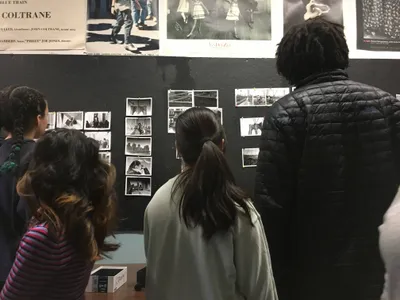 Students studying contact sheets and prints pinned to a dark photography studio wall, below posters of jazz and fashion photographs
