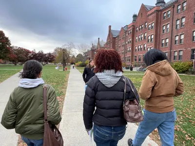 Students following a guide along a path through a college campus on an autumn visit, brick halls to one side and fallen leaves on the grass