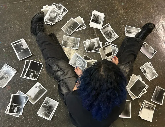 A student sitting on a studio floor surrounded by dozens of black-and-white prints spread out around them, holding one up to examine it