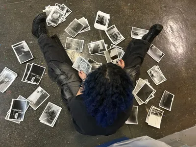 A student sitting on a studio floor surrounded by dozens of black-and-white prints spread out around them, holding one up to examine it