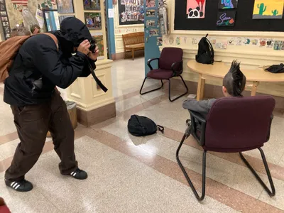A student crouching to photograph a classmate seated on a chair in the school lobby, setting up a portrait shot