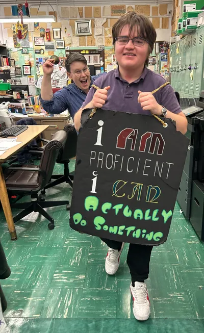 A student grinning as he holds up a hand-lettered sign reading "I am proficient, I can actually do something", with a teacher cheering behind him in the staff room