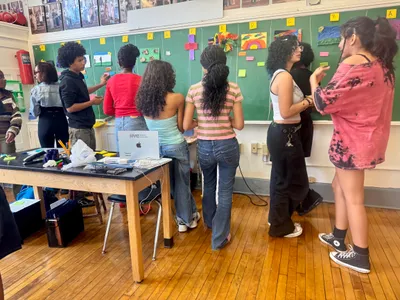 Students standing along a green chalkboard hung with lettered artworks, adding sticky notes as they move down the row in a gallery-walk critique