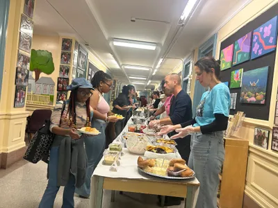 Students and staff serving themselves from a long buffet table set up in a school corridor lined with framed photographs and student paintings