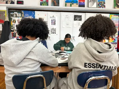 Two students seen from behind working at a table while a classmate opposite leafs through a large book of images