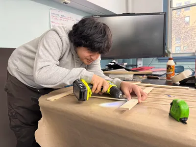 A student using a cordless drill to fasten together a wooden frame on a paper-covered worktable
