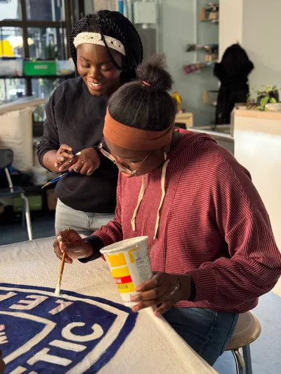 Two students painting a large fabric banner spread across a table, one holding a tub of paint while the other works with a brush