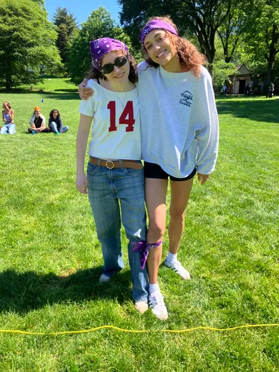 Two students in matching purple bandanas standing arm in arm on a sunny lawn during a field day, their legs tied together for a three-legged race
