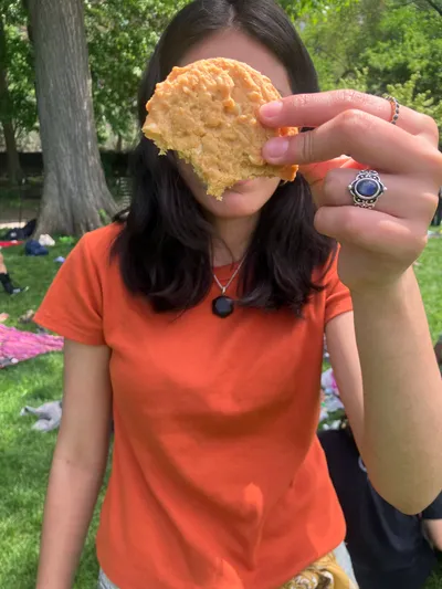 A student holding a large round cracker up in front of her face while sitting on the grass in a park