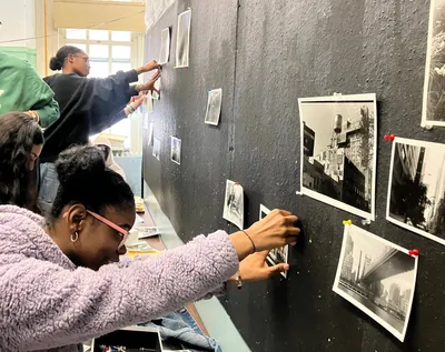 Two students pinning their black-and-white photographic prints onto a dark studio wall for a critique