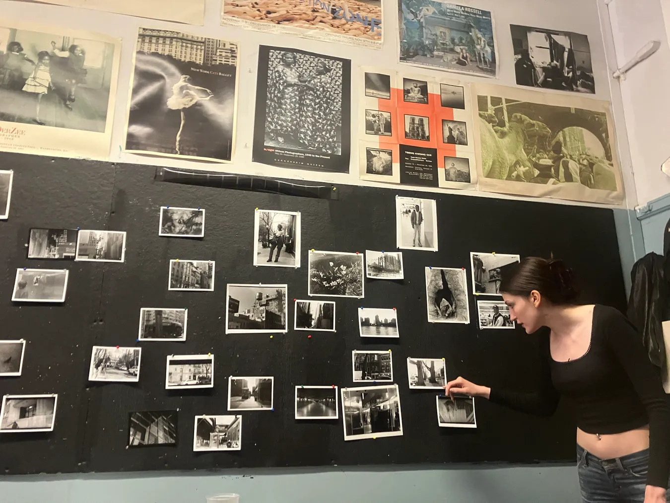 A student pinning her black-and-white prints to a dark studio wall already covered with photographs, beneath a row of exhibition posters