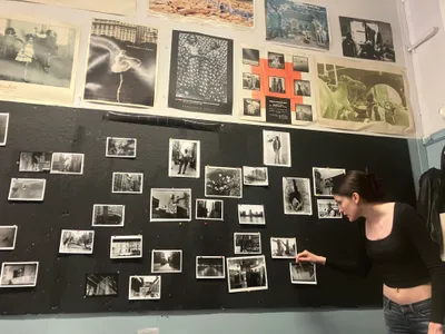 A student pinning her black-and-white prints to a dark studio wall already covered with photographs, beneath a row of exhibition posters