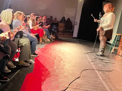 A student standing at a microphone on a small stage, presenting to classmates who sit along the front row following printed copies of the work