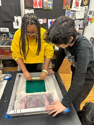 Two students pulling a squeegee of green ink across a screen-printing frame on a studio table, the wall behind them covered in prints and posters