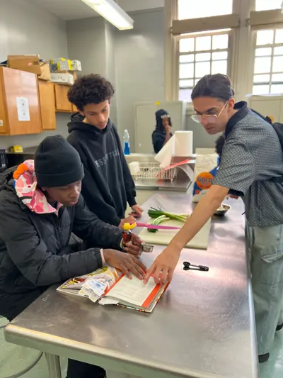 Students gathered around a stainless steel counter in the school kitchen, working through a recipe on a clipboard together