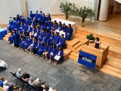 Graduates in blue caps and gowns seated on tiered steps while a student speaks at the lectern beside an Urban Academy sign, families watching from the floor below