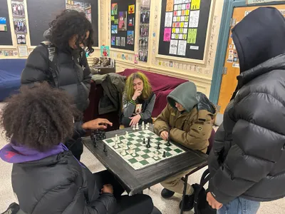 Students playing a game of chess at a small table in a common room, with classmates gathered around to watch