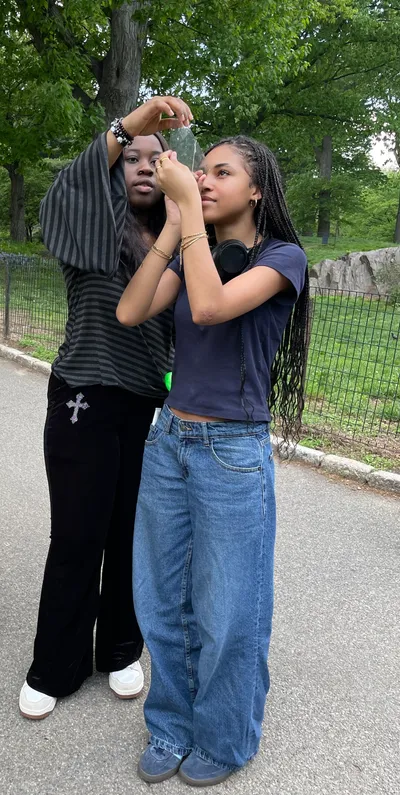 Two students standing on a park path, one holding up a small mirror while the other lines up a photograph through it