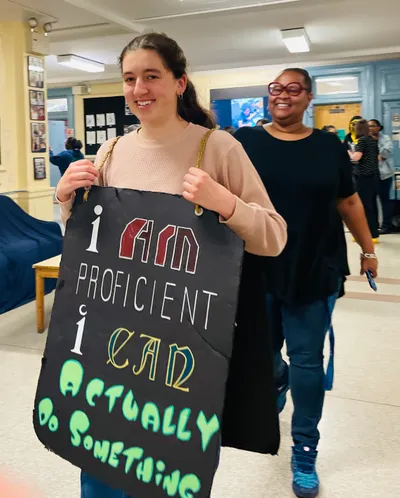 A student wearing a hand-lettered sandwich board reading "I am proficient, I can actually do something" walking down a school corridor, a teacher smiling behind her