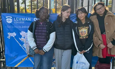 Four students standing in front of a blue Lehman College banner during a campus visit, one holding a Lehman College tote bag
