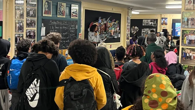 A packed room of students standing with their backpacks, listening to a classmate speaking at the front beneath a chalkboard and a wall covered in photographs