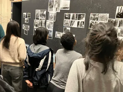 Students standing in front of a black display wall pinned with dozens of black-and-white photographs, studying the work on show