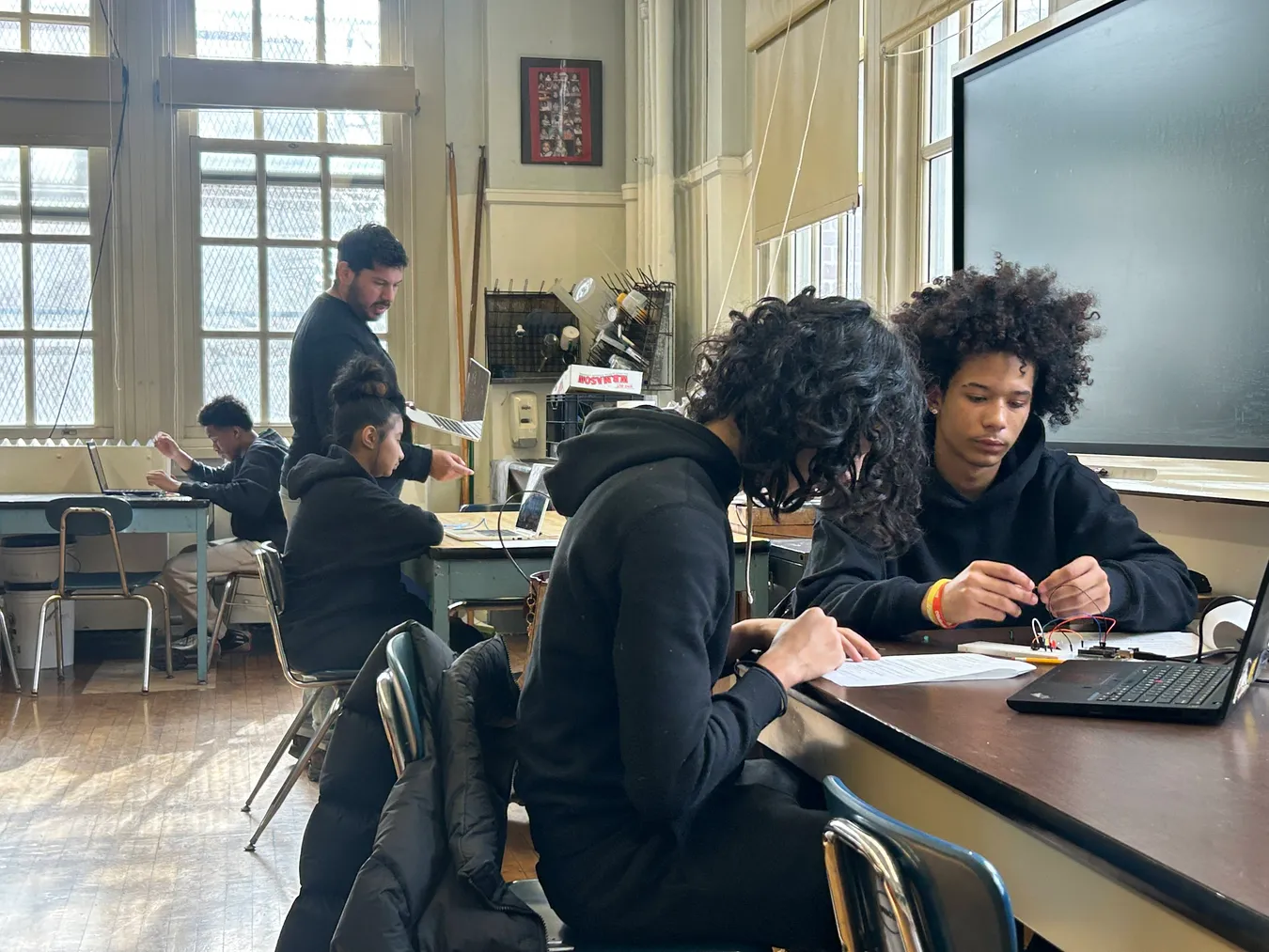 Students working in pairs at tables in a science room, one examining a small wired component while a teacher helps another group by the windows