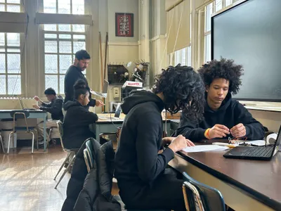 Students working in pairs at tables in a science room, one examining a small wired component while a teacher helps another group by the windows
