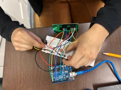A student's hands running jumper wires between a breadboard, a sensor module and an Arduino board on a desk