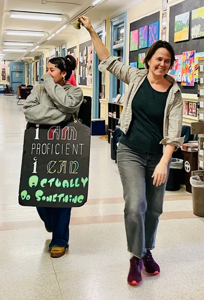 A student laughing behind her hand as she walks a corridor hung with student paintings, wearing a hand-lettered sandwich board reading "I am proficient, I can actually do something", while a teacher rings a brass bell overhead beside her