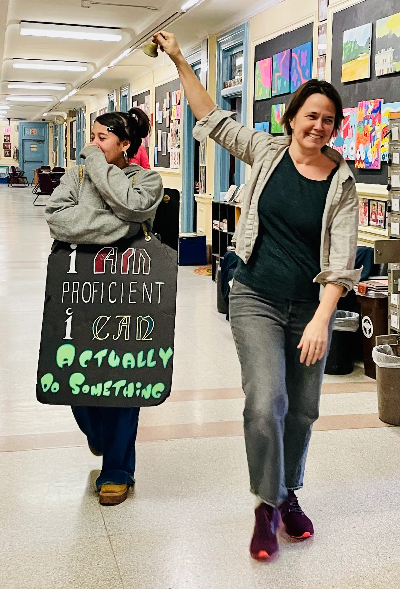 A student laughing behind her hand as she walks a corridor hung with student paintings, wearing a hand-lettered sandwich board reading "I am proficient, I can actually do something", while a teacher rings a brass bell overhead beside her