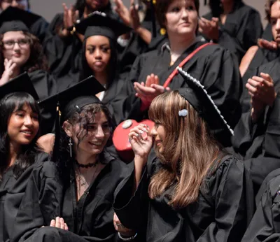 Two graduates in caps and gowns laughing together while classmates applaud around them at the graduation ceremony