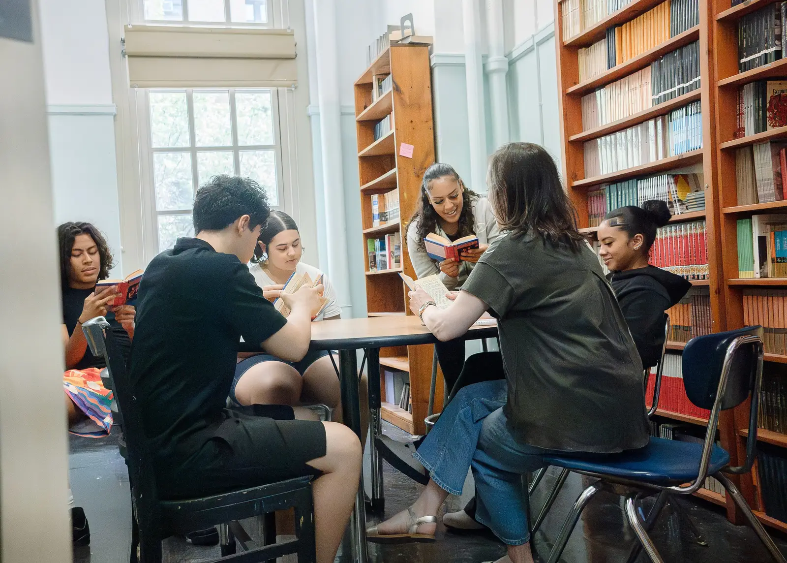 Six students and a teacher sitting in a circle of chairs in the library, each holding an open book, surrounded by full bookshelves