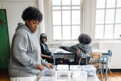 A student sorting hardware into labelled plastic tubs at a table in a bright classroom with tall windows, classmates working behind her