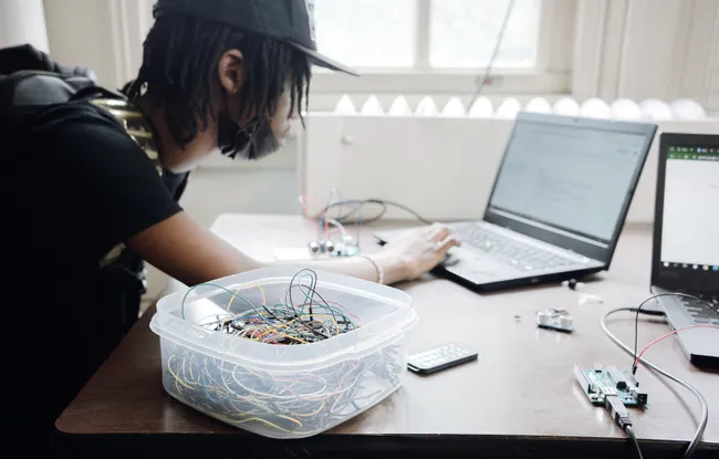 A student leaning over a laptop to program a small microcontroller board, with a tub of tangled jumper wires open on the desk beside them