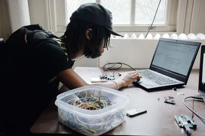 A student leaning over a laptop to program a small sensor circuit, with a tub of jumper wires on the desk beside them