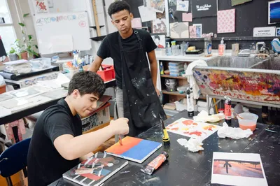A student painting a canvas in bright orange and blue while a classmate in a painting apron looks on, in a busy art studio with a paint-spattered sink behind them