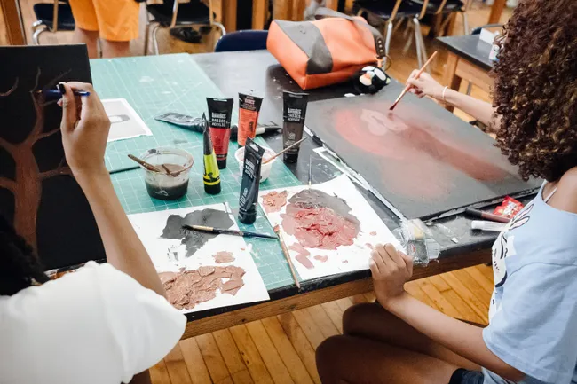 Two students painting at a shared studio table covered in acrylic tubes and mixed paint, one working on a tree silhouette and the other on a soft red abstract canvas