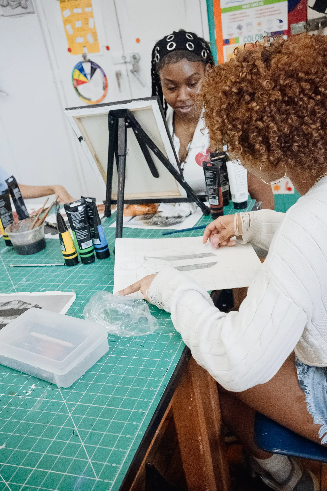 Two students at a cutting mat covered in paint tubes and brushes, looking together at a pencil drawing, with a canvas on an easel behind them