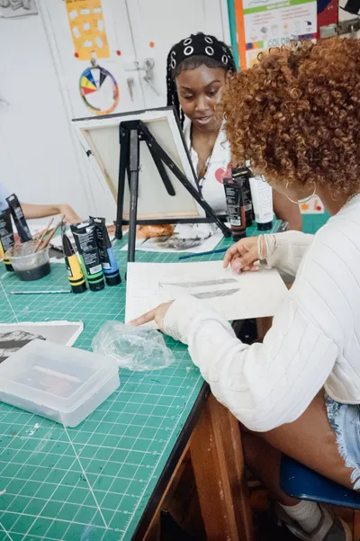 Two students at a cutting mat covered in paint tubes and brushes, looking together at a pencil drawing, with a canvas on an easel behind them
