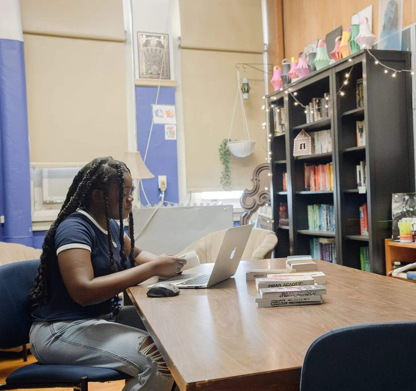 A student reading at a long table in the school library, laptop open in front of her and a stack of bound Urban Academy project books beside her