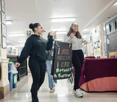 Three students walking down a school corridor, laughing, one carrying a hand-lettered sign reading "I am proficient, I can actually do something"