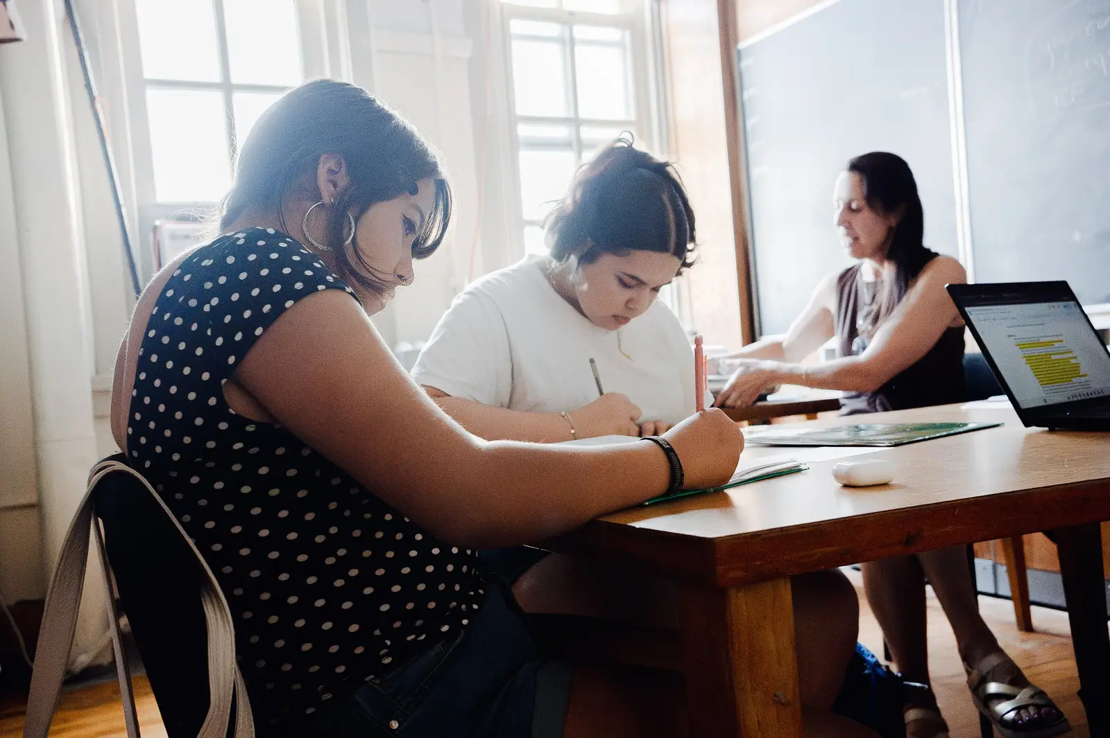 Two students bent over notebooks writing at a sunlit table, a teacher helping another student at a laptop showing a highlighted document in front of a chalkboard behind them